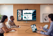 Dell simplifica las videoconferencias con un monitor 4K todo en uno Fotografía de tres profesionales en una sala de reuniones pequeña, participando en una videoconferencia a través de un monitor Dell de gran tamaño montado en una estructura de madera. La pantalla, conectada a una computadora portátil sobre la mesa mediante un único cable negro, está dividida mostrando a cuatro participantes remotos en el lado izquierdo y un panel con gráficos de datos en el derecho.