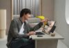 ASUS impulsa la productividad con laptops de doble pantalla Fotografía de un hombre joven sonriente utilizando una laptop ASUS Zenbook DUO sobre un escritorio minimalista con acabado de mármol. El equipo tiene sus dos pantallas desplegadas verticalmente, mostrando una galería de imágenes y herramientas de diseño. El usuario toca la pantalla superior con una mano mientras utiliza el teclado magnético separado con la otra.