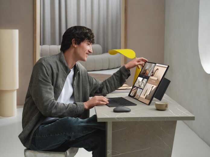 Fotografía de un hombre joven sonriente utilizando una laptop ASUS Zenbook DUO sobre un escritorio minimalista con acabado de mármol. El equipo tiene sus dos pantallas desplegadas verticalmente, mostrando una galería de imágenes y herramientas de diseño. El usuario toca la pantalla superior con una mano mientras utiliza el teclado magnético separado con la otra.