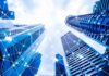 Schneider Electric eleva su perspectiva para 2026 tras semestre récord impulsado por los centros de datos Fotografía en ángulo contrapicado de rascacielos corporativos de cristal apuntando hacia un cielo parcialmente nublado. Sobre los edificios se superpone una red digital de líneas interconectadas y nodos brillantes que simbolizan la conectividad global, la transferencia de datos y las redes de energía inteligente.