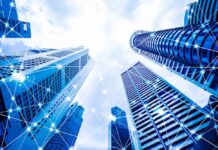 Schneider Electric eleva su perspectiva para 2026 tras semestre récord impulsado por los centros de datos Fotografía en ángulo contrapicado de rascacielos corporativos de cristal apuntando hacia un cielo parcialmente nublado. Sobre los edificios se superpone una red digital de líneas interconectadas y nodos brillantes que simbolizan la conectividad global, la transferencia de datos y las redes de energía inteligente.