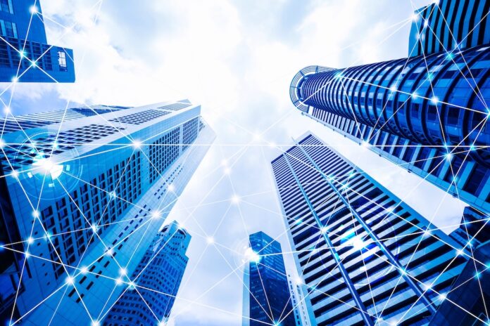 Fotografía en ángulo contrapicado de rascacielos corporativos de cristal apuntando hacia un cielo parcialmente nublado. Sobre los edificios se superpone una red digital de líneas interconectadas y nodos brillantes que simbolizan la conectividad global, la transferencia de datos y las redes de energía inteligente.