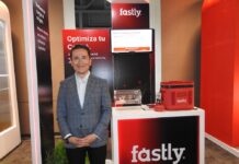 Fastly redefine la seguridad en el edge y abre nuevas oportunidades de crecimiento Fotografía de Ricardo Caballero, country manager de México y Centroamérica para Fastly. Viste un saco gris a cuadros y posa sonriente frente al stand de su compañía, iluminado con tonos rojizos. El módulo promocional tiene un monitor con una presentación, el logotipo de la empresa, un contenedor de acrílico y obsequios.