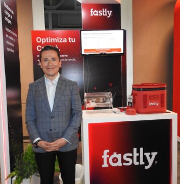 Fastly redefine la seguridad en el edge y abre nuevas oportunidades de crecimiento Fotografía de Ricardo Caballero, country manager de México y Centroamérica para Fastly. Viste un saco gris a cuadros y posa sonriente frente al stand de su compañía, iluminado con tonos rojizos. El módulo promocional tiene un monitor con una presentación, el logotipo de la empresa, un contenedor de acrílico y obsequios.