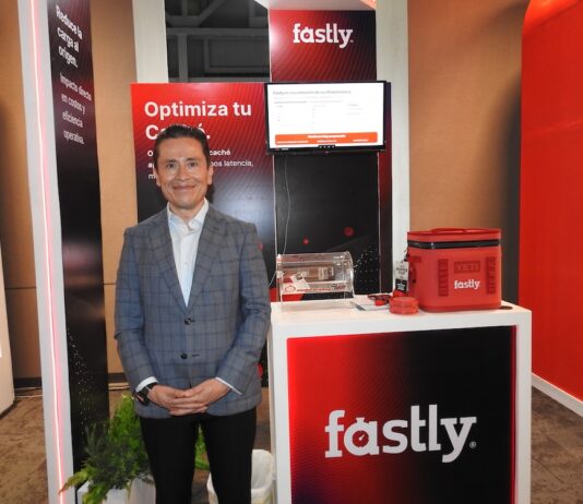 Fastly redefine la seguridad en el edge y abre nuevas oportunidades de crecimiento Fotografía de Ricardo Caballero, country manager de México y Centroamérica para Fastly. Viste un saco gris a cuadros y posa sonriente frente al stand de su compañía, iluminado con tonos rojizos. El módulo promocional tiene un monitor con una presentación, el logotipo de la empresa, un contenedor de acrílico y obsequios.