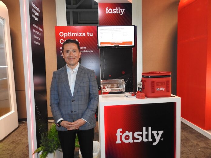Seguridad y rendimiento procesados en el borde Fotografía de Ricardo Caballero, country manager de México y Centroamérica para Fastly. Viste un saco gris a cuadros y posa sonriente frente al stand de su compañía, iluminado con tonos rojizos. El módulo promocional tiene un monitor con una presentación, el logotipo de la empresa, un contenedor de acrílico y obsequios.