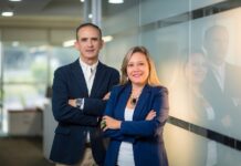 Hitachi Vantara fortalece al canal con soluciones de ciberresiliencia Fotografía corporativa de César Zurita y Claudia Aro, directivos de Hitachi Vantara, posando de pie y con los brazos cruzados en un pasillo de oficina moderno. Ambos visten sacos de color azul oscuro y se encuentran junto a una pared de cristal esmerilado que refleja la iluminación cálida del entorno.