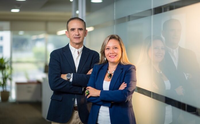 Fotografía corporativa de César Zurita y Claudia Aro, directivos de Hitachi Vantara, posando de pie y con los brazos cruzados en un pasillo de oficina moderno. Ambos visten sacos de color azul oscuro y se encuentran junto a una pared de cristal esmerilado que refleja la iluminación cálida del entorno.