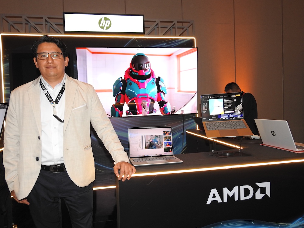 Fotografía de un hombre con anteojos y saco claro de pie frente a un stand iluminado de HP y AMD. En el mostrador se exhiben computadoras portátiles en funcionamiento y, al fondo, una pantalla grande muestra el modelado 3D de un personaje con armadura robótica colorida.