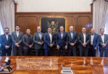 Schneider Electric amplía su manufactura en Querétaro con tres nuevas plantas Fotografía grupal de diez hombres con traje, que incluye a funcionarios del gobierno de Querétaro y directivos de Schneider Electric, posando de pie y sonriendo frente a un muro de madera adornado con el escudo oficial del estado.