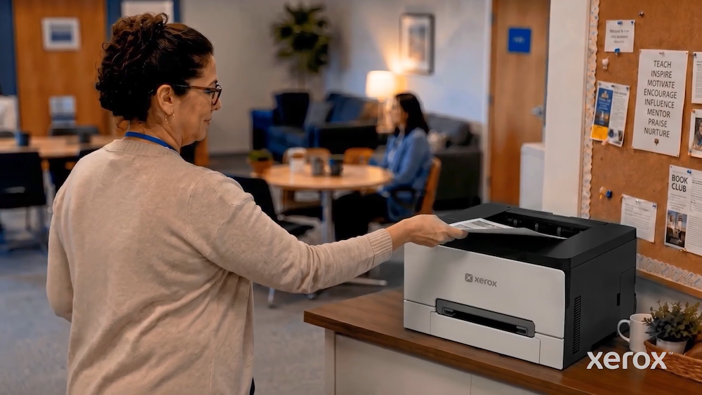 Una mujer en un entorno de oficina casual o sala de maestros recogiendo una hoja de papel recién impresa de una impresora de escritorio Xerox color blanco y negro, ubicada sobre un mueble de madera junto a un tablero de anuncios.