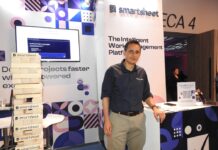 Systec Consulting impulsa la adopción de Smartsheet en América Latina Fotografía de Jorge Díaz, CEO de Systec Consulting, de pie en un módulo de exhibición de Smartsheet. A su izquierda se encuentra una torre gigante de bloques tipo Jenga de madera, donde cada pieza tiene impreso el logotipo de la marca de software.