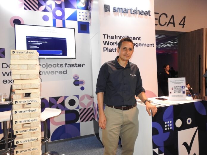 Fotografía de Jorge Díaz, CEO de Systec Consulting, de pie en un módulo de exhibición de Smartsheet. A su izquierda se encuentra una torre gigante de bloques tipo Jenga de madera, donde cada pieza tiene impreso el logotipo de la marca de software.