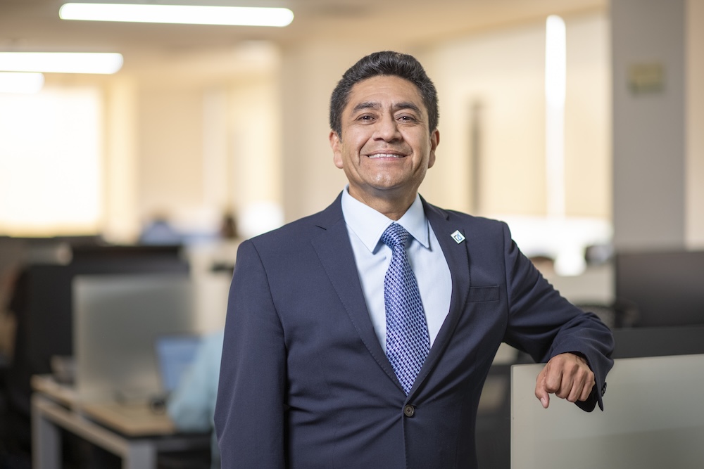 Fotografía de Ángel Baños Carrillo sonriendo a la cámara. Viste un traje azul marino con una corbata azul de patrón geométrico y camisa clara, y se encuentra apoyado ligeramente sobre una mampara de cristal en un entorno de oficina.