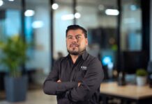 VIGI Cloud VMS simplifica la videovigilancia y elimina hardware dedicado Fotografía de medio cuerpo de Armando Hernández, Channel Sales Manager de TP-Link México, posando con los brazos cruzados y vistiendo una camisa negra con el logotipo de VIGI by TP-Link, con un fondo de oficina desenfocado.