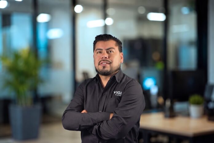 Fotografía de medio cuerpo de Armando Hernández, Channel Sales Manager de TP-Link México, posando con los brazos cruzados y vistiendo una camisa negra con el logotipo de VIGI by TP-Link, con un fondo de oficina desenfocado.