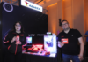 Adata revoluciona el rendimiento extremo para los entusiastas del hardware Fotografía en plano medio de Adriana Morales y Donato Moreno en el estand de ADATA y XPG, sosteniendo empaques de memorias RAM y unidades SSD de quinta generación junto a un gabinete de computadora transparente iluminado con luces LED RGB y periféricos.