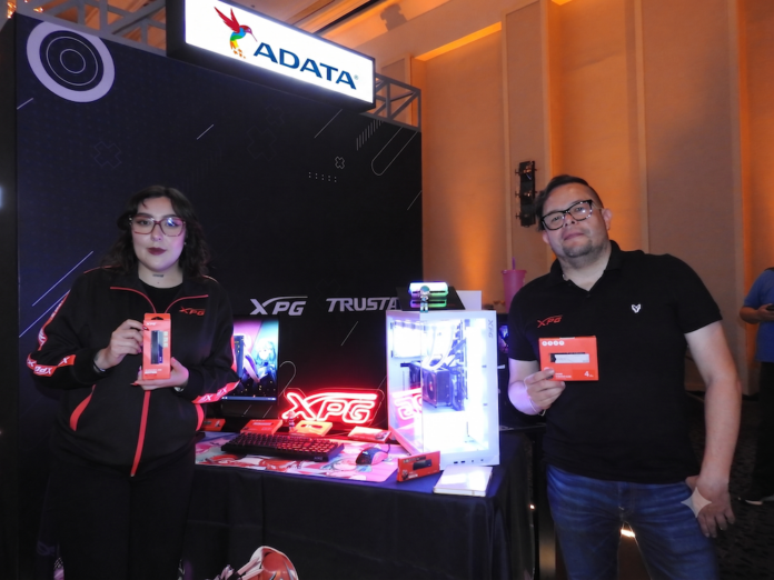 Fotografía en plano medio de Adriana Morales y Donato Moreno en el estand de ADATA y XPG, sosteniendo empaques de memorias RAM y unidades SSD de quinta generación junto a un gabinete de computadora transparente iluminado con luces LED RGB y periféricos.
