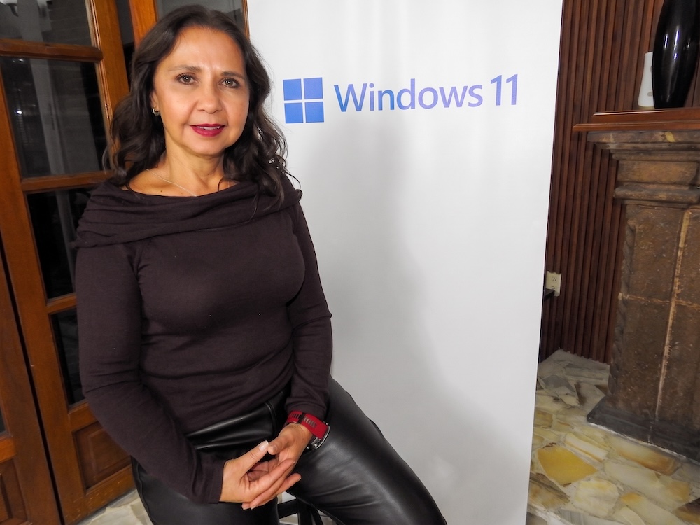 Fotografía de retrato de Adriana Valdez sentada en un taburete frente a una mampara blanca con el logotipo de Windows 11. Viste una blusa de cuello drapeado color café oscuro y pantalones de piel negros.