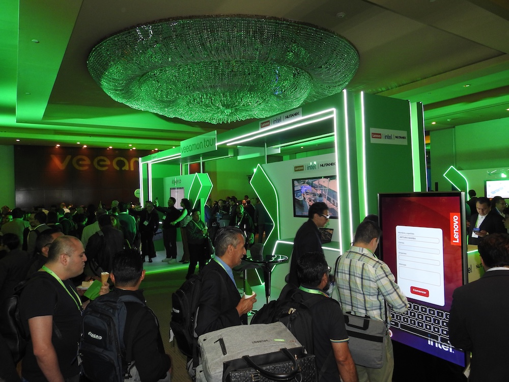 Salón de exposiciones iluminado con luces verdes lleno de asistentes interactuando en los booths del VeeamOn Tour.