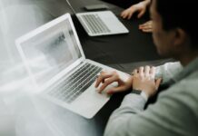 Akamai advierte sobre el aumento de ciberriesgos por el uso de IA en las empresas Fotografía en ángulo picado de un empleado trabajando con una computadora portátil plateada sobre una mesa oscura dentro de una oficina, utilizando el trackpad con su mano derecha mientras otro colaborador utiliza una laptop en segundo plano.