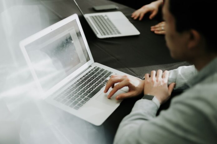 Fotografía en ángulo picado de un empleado trabajando con una computadora portátil plateada sobre una mesa oscura dentro de una oficina, utilizando el trackpad con su mano derecha mientras otro colaborador utiliza una laptop en segundo plano.