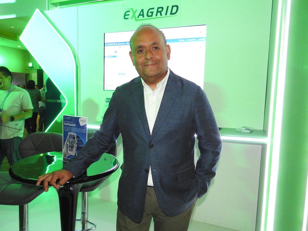 Ejecutivo sonriendo en el stand de ExaGrid con saco azul marino y el fondo verde de la marca.