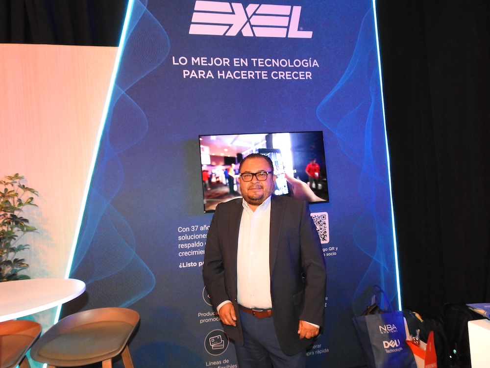 Fotografía en plano medio de Amador Rangel, Gerente de Dell Cómputo en Exel del Norte, luciendo un saco oscuro y anteojos frente a un panel publicitario retroiluminado de la compañía que muestra el lema "LO MEJOR EN TECNOLOGÍA PARA HACERTE CRECER".