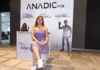 Impulso empresarial femenino 2026: Liderazgo y redes de negocio Anadic Mx Fotografía de Sonia Rodríguez Cansino sentada con las piernas cruzadas sobre un taburete alto en un pabellón de exposiciones. Viste un vestido corto de color morado claro y tenis blancos. A su espalda se observa un gran panel publicitario con el logotipo de ANADIC MX y sus comités como "Mujeres ANADIC MX" y "Jóvenes ANADIC MX", además de un pendón de "IT Leaders Hub" a la derecha.