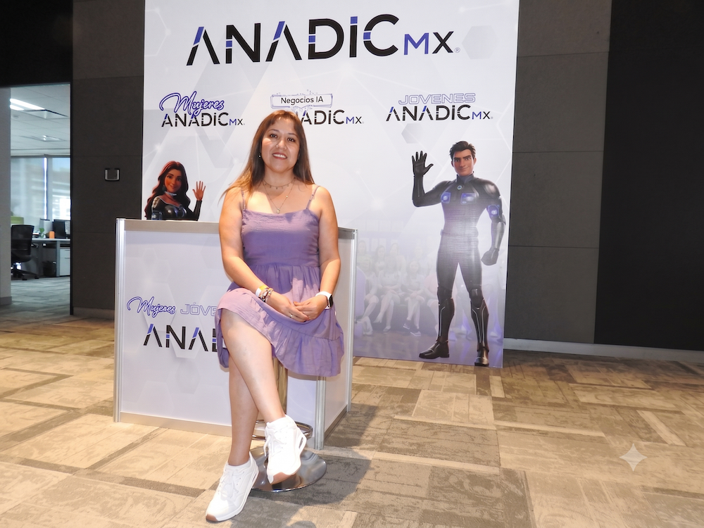 Anadic Mx consolida el liderazgo ejecutivo femenino en el sector de las TICs Fotografía de Sonia Rodríguez Cancino sentada con las piernas cruzadas sobre un taburete alto en un pabellón de exposiciones. Viste un vestido corto de color morado claro y tenis blancos. A su espalda se observa un gran panel publicitario con el logotipo de ANADIC MX y sus comités como