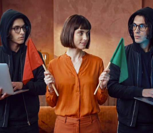 Avast revela que el 53% de los mexicanos cree detectar contenido falso pero el 65% expone sus datos a chatbots de IA Concepto visual en un entorno de oficina donde una mujer vestida con blusa naranja se encuentra entre dos figuras idénticas con sudadera negra con capucha y anteojos sosteniendo laptops; la mujer sostiene una bandera roja hacia la izquierda y una bandera verde hacia la derecha para ejemplificar la toma de decisiones frente a señales de alerta digital.