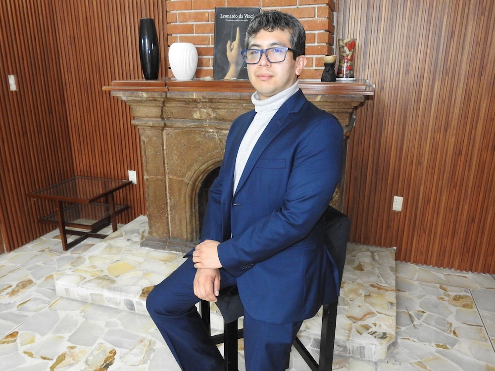 Fotografía de retrato de Carlos Enrique García sentado de perfil sobre un taburete, vistiendo traje azul marino con suéter de cuello alto gris y anteojos, posando frente a una chimenea de piedra.