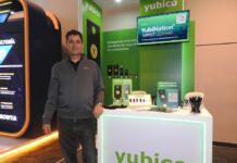 Autenticación multifactor sin contraseña: Yubico refuerza la ciberseguridad en 2026 Fotografía de Juan Ignacio Quesada posando de pie con la mano apoyada sobre el mostrador de exhibición iluminado de Yubico. Viste un suéter gris con el logotipo de la marca y anteojos. Detrás de él se observa una pared verde con pantallas corporativas, folletos informativos, muestras físicas de llaves de seguridad YubiKey y un monitor promocional.