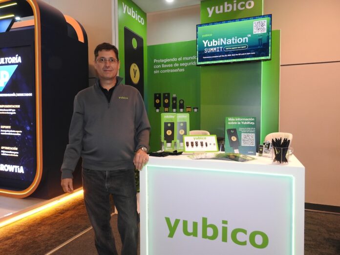 Fotografía de Juan Ignacio Quesada posando de pie con la mano apoyada sobre el mostrador de exhibición iluminado de Yubico. Viste un suéter gris con el logotipo de la marca y anteojos. Detrás de él se observa una pared verde con pantallas corporativas, folletos informativos, muestras físicas de llaves de seguridad YubiKey y un monitor promocional.