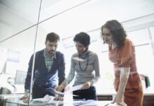 Dxgrow acelera el desarrollo de talento y eleva la productividad en consultoría SAP con inteligencia artificial Fotografía de tres consultores —un hombre y dos mujeres— reunidos de pie frente a una mesa de cristal en una oficina iluminada, revisando conjuntamente documentos y gráficos impresos.
