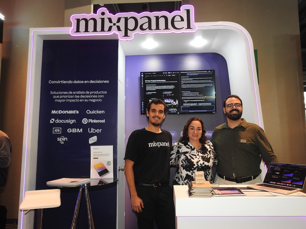 Fotografía de tres personas, una mujer en el centro y dos hombres a los costados, sonriendo a la cámara desde el mostrador de exhibición de Mixpanel en un evento tecnológico. El módulo cuenta con computadoras portátiles, libretas promocionales y una pantalla superior que muestra herramientas de métricas interactivas.