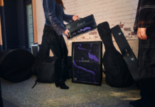 Marshall celebra a Jimi Hendrix con nuevas ediciones por su 60 aniversario Fotografía de una persona sosteniendo el cabezal del amplificador JMH Studio de Marshall sobre su respectivo gabinete en la calle. Los equipos destacan por su patrón psicodélico morado y negro, y están rodeados de diversos estuches rígidos para instrumentos musicales.