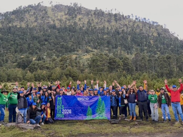 Fotografía grupal que reúne a decenas de voluntarios sonrientes alzando los brazos en una pradera al pie de una montaña arbolada, sosteniendo una lona azul con los logotipos de Epson y la leyenda 