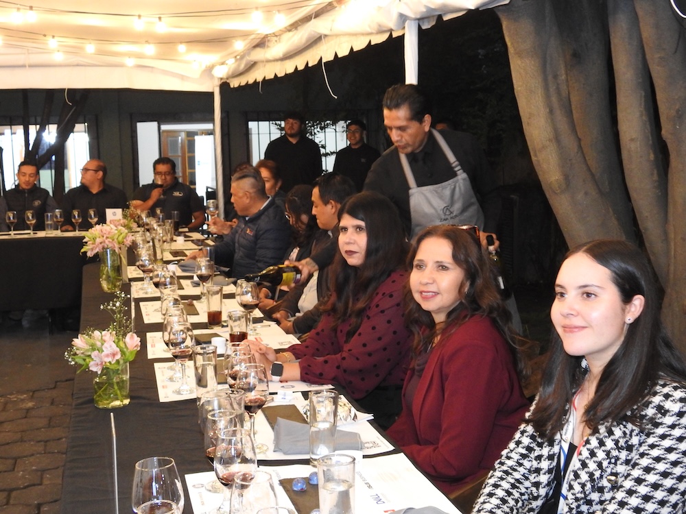 Fotografía en ángulo diagonal de una mesa larga servida para un maridaje, donde un mesero sirve vino tinto en la copa de una ejecutiva. Las invitadas conversan bajo un techo con guirnaldas de luces cálidas.