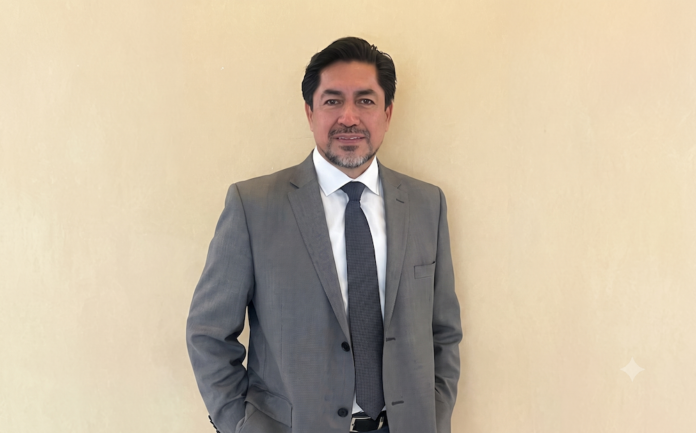 Estrategia y resiliencia para el fortalecimiento del canal en ciberseguridad Fotografía de retrato de Fausto Escobar posando de pie con las manos en los bolsillos, vistiendo un traje gris, camisa blanca y corbata azul oscuro, sobre un fondo plano de color beige claro.