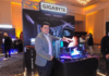 Gigabyte impulsa la gama media ante el nuevo escenario de precios Fotografía de Jhon Restrepo, Marketing Manager de Gigabyte, vistiendo un saco gris y camisa clara, señalando un ecosistema de cómputo exhibido sobre una mesa negra. El montaje incluye un monitor, teclado, mouse, gabinete de PC y tarjeta gráfica, ubicados frente a un stand iluminado con los logotipos de AMD y Gigabyte bajo el lema "Experiencia Única". Al fondo se observan asistentes recorriendo el evento corporativo.