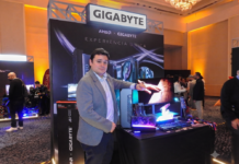 Gigabyte impulsa la gama media ante el nuevo escenario de precios Fotografía de Jhon Restrepo, Marketing Manager de Gigabyte, vistiendo un saco gris y camisa clara, señalando un ecosistema de cómputo exhibido sobre una mesa negra. El montaje incluye un monitor, teclado, mouse, gabinete de PC y tarjeta gráfica, ubicados frente a un stand iluminado con los logotipos de AMD y Gigabyte bajo el lema "Experiencia Única". Al fondo se observan asistentes recorriendo el evento corporativo.