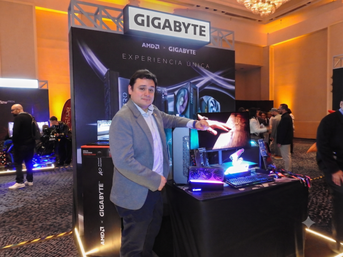 Expansión estratégica de Gigabyte en el segmento de gama media Fotografía de Jhon Restrepo, Marketing Manager de Gigabyte, vistiendo un saco gris y camisa clara, señalando un ecosistema de cómputo exhibido sobre una mesa negra. El montaje incluye un monitor, teclado, mouse, gabinete de PC y tarjeta gráfica, ubicados frente a un stand iluminado con los logotipos de AMD y Gigabyte bajo el lema