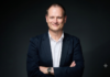 Hitachi Vantara: ¿Quién controla los datos? La pregunta que los directorios ya no pueden ignorar en la era de la IA Fotografía de retrato de Adrian Johnson, sonriendo ligeramente con los brazos cruzados, vistiendo un saco azul marino sobre una camisa blanca de cuello abierto, frente a un fondo neutro de color oscuro.