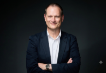 Hitachi Vantara: ¿Quién controla los datos? La pregunta que los directorios ya no pueden ignorar en la era de la IA Fotografía de retrato de Adrian Johnson, sonriendo ligeramente con los brazos cruzados, vistiendo un saco azul marino sobre una camisa blanca de cuello abierto, frente a un fondo neutro de color oscuro.