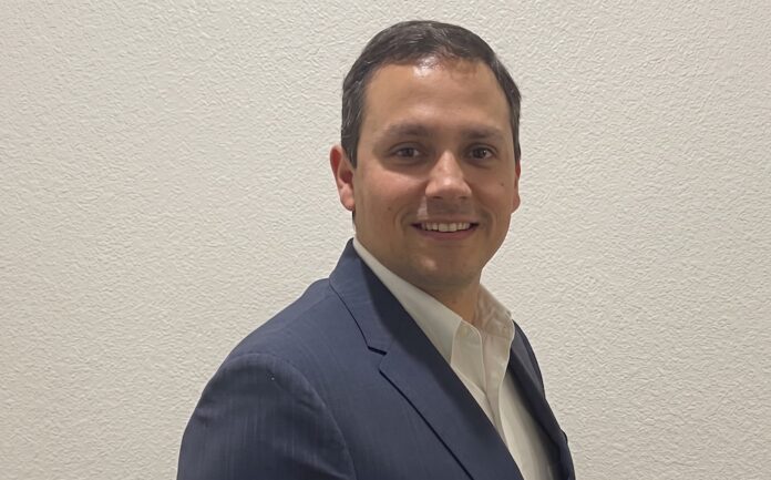Fotografía de retrato de Heriberto Cabrera posando de tres cuartos con una sonrisa sutil frente a una pared de fondo claro. Viste un saco azul marino formal sobre una camisa blanca de cuello abierto.