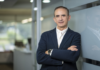 Hitachi Vantara nombra a César Zurita como nuevo Director Comercial en México Fotografía de retrato ejecutivo de César Zurita posando de tres cuartos con los brazos cruzados, apoyado sutilmente sobre un panel de cristal en una oficina moderna. Viste un saco de vestir color azul marino sobre una camisa blanca de cuello abierto y porta un reloj inteligente en la muñeca izquierda.