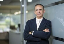 Hitachi Vantara nombra a César Zurita como nuevo Director Comercial en México Fotografía de retrato ejecutivo de César Zurita posando de tres cuartos con los brazos cruzados, apoyado sutilmente sobre un panel de cristal en una oficina moderna. Viste un saco de vestir color azul marino sobre una camisa blanca de cuello abierto y porta un reloj inteligente en la muñeca izquierda.