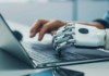 Hostinger impulsa la evolución del ecommerce en México frente al avance de la IA Fotografía de concepto tecnológico que muestra en primer plano una mano robótica articulada de color blanco escribiendo sobre el teclado de una computadora portátil plateada.