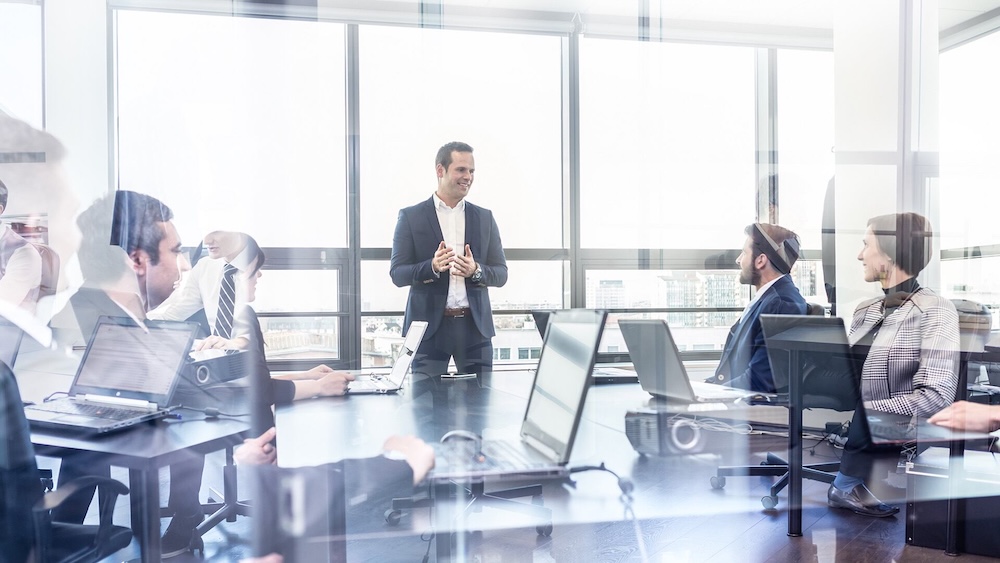 Fotografía capturada a través de un cristal corporativo que muestra a un ejecutivo hablando de pie frente a un equipo de trabajo sentado en una sala de reuniones bien iluminada con computadoras portátiles sobre la mesa.